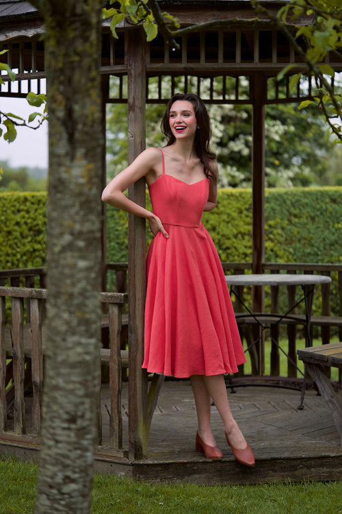 A girl standing against the backdrop of a castle, wearing a Brigitte Bardot inspired red linen dress as a guest to a wedding. 