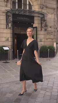 A blonde standing in front of a hotel in Paris wearing a long black evening dress from Gaâla