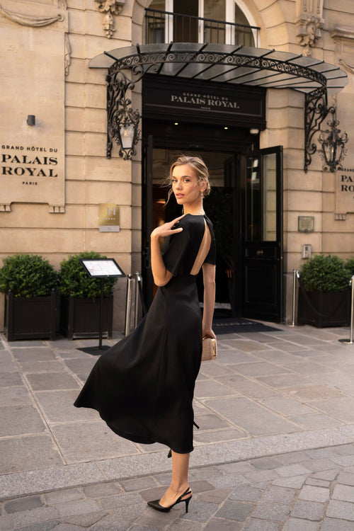 A blonde girl standing in front of a fancy hotel wearing a long black evening dress from Gaâla