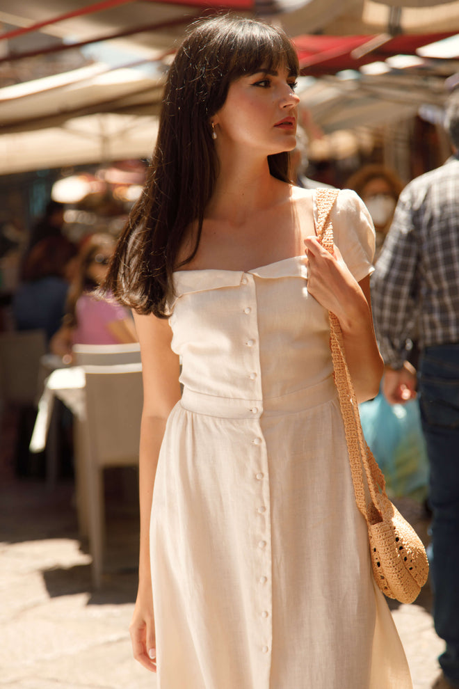 Brunette girl in a marketplace in Sicily, wearing a vintage inspired button down cream Gaâla linen dress