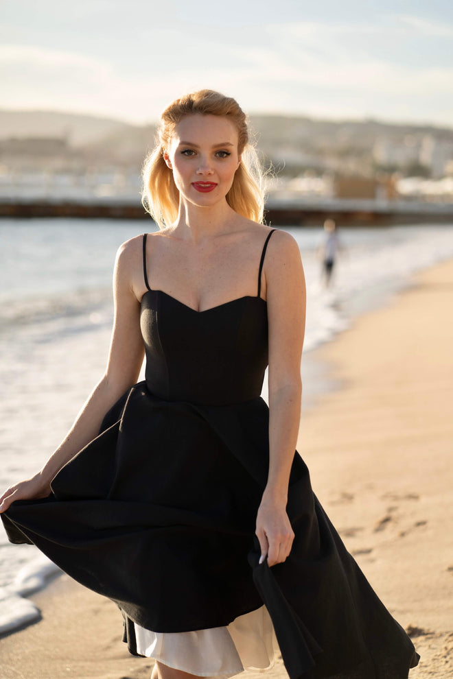 A blonde girl recreating Brigitte Bardot’s iconic run on the beach in Cannes, wearing a Gaâla cotton/linen black Bardot dress.